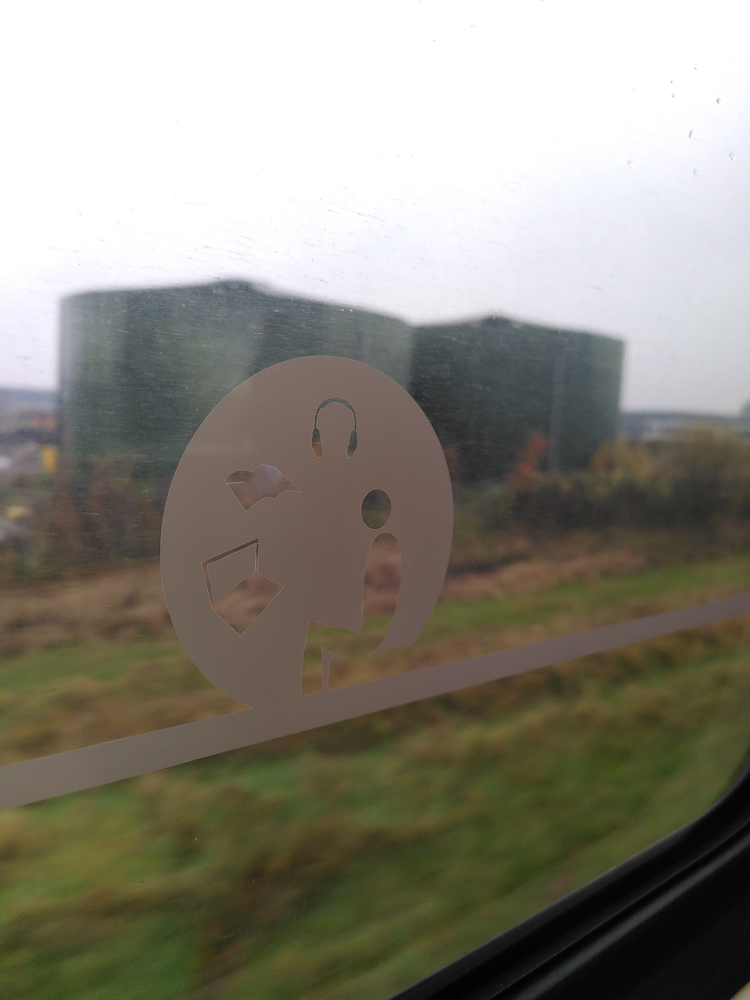 A round white sign on a train window. There's an armless person sitting. What is possibly a laptop, probably a book, and a pair of headphones floating near by. Outside the window is a blurry boxy building